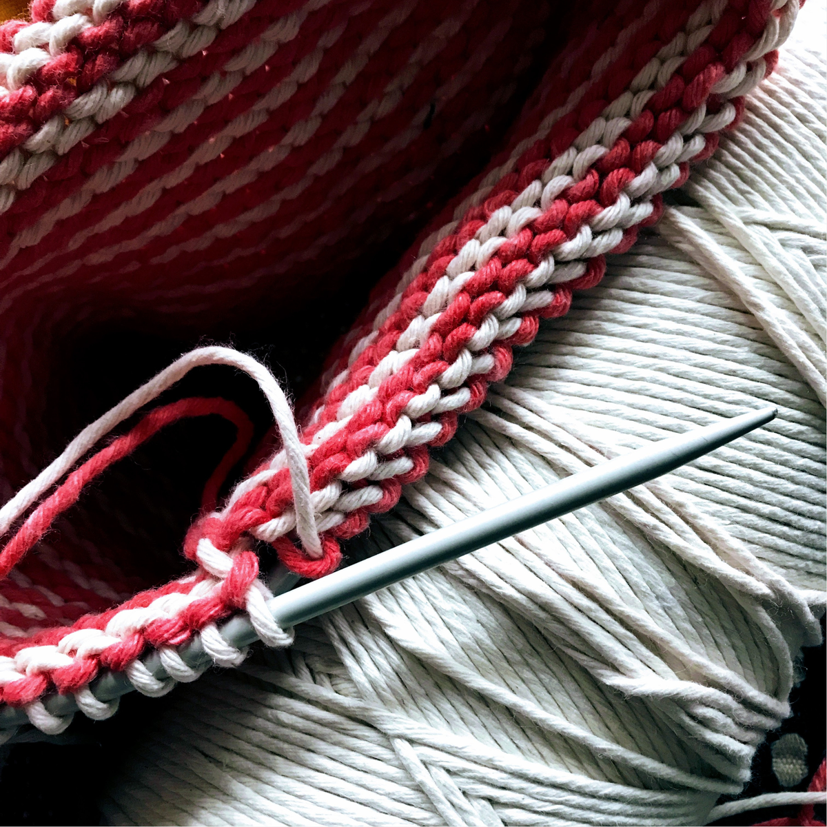 Close-up of red and white yarn being knitted on circular needles against a spool of cotton string