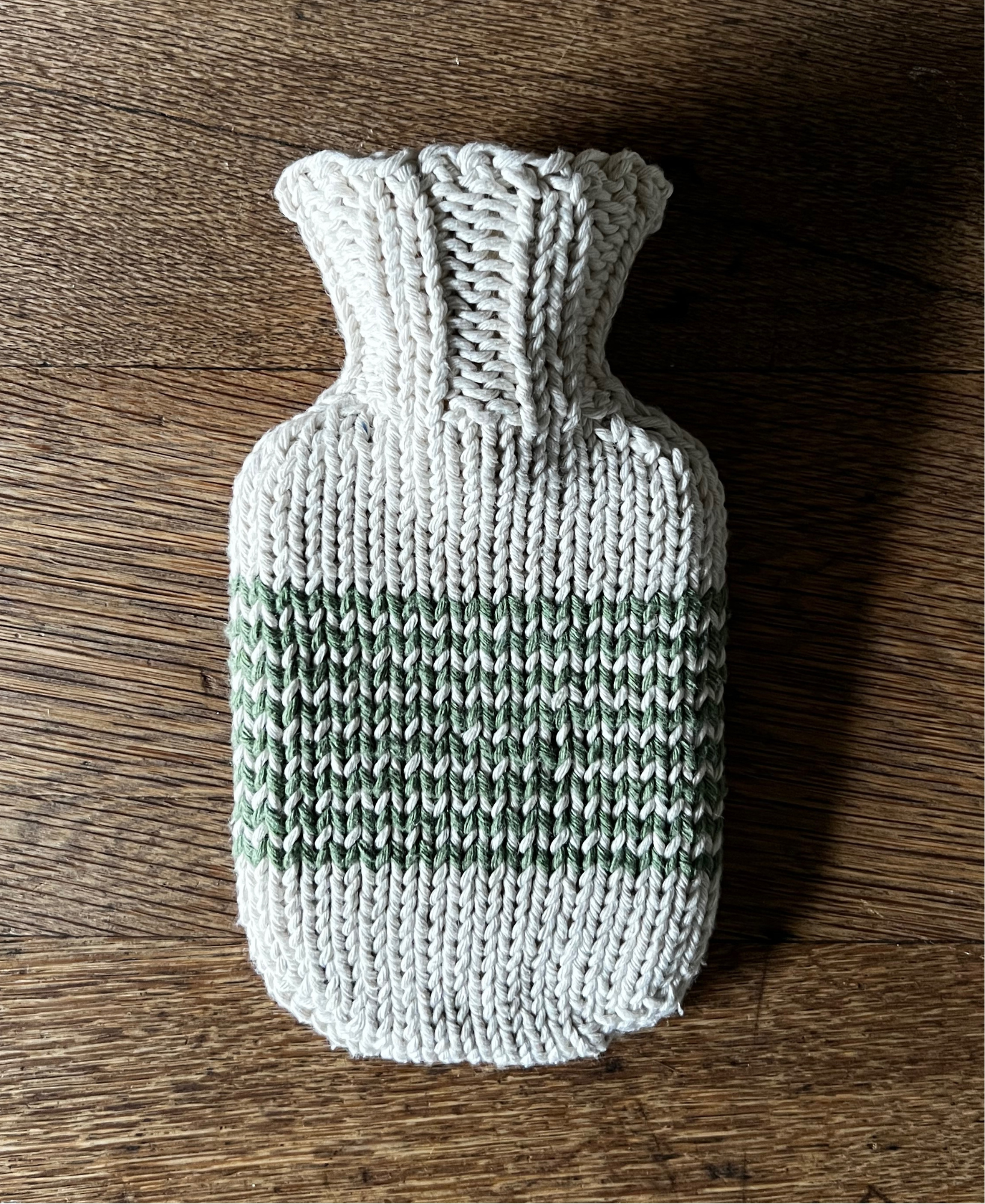 Knitted hot water bottle cover with a green stripe on a wooden surface