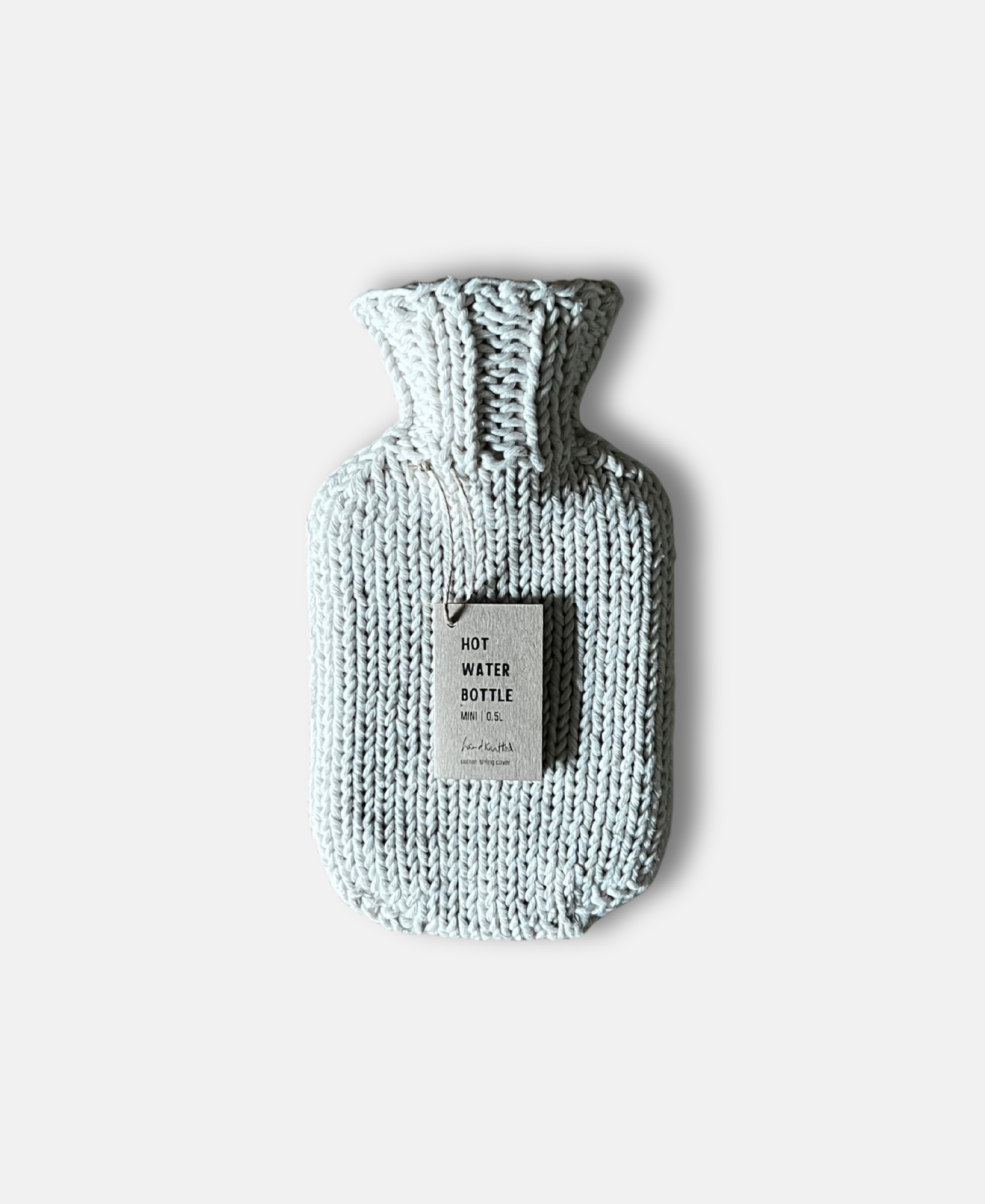 Knitted cotton string hot water bottle with kraft tag on a pale grey background