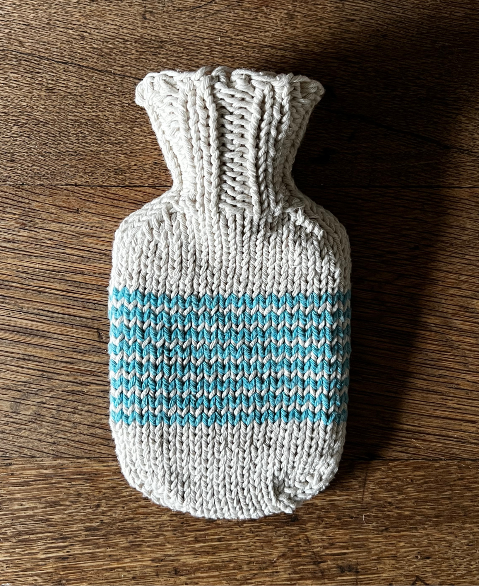 Knitted hot water bottle cover with turquoise stripe placed on a wooden surface