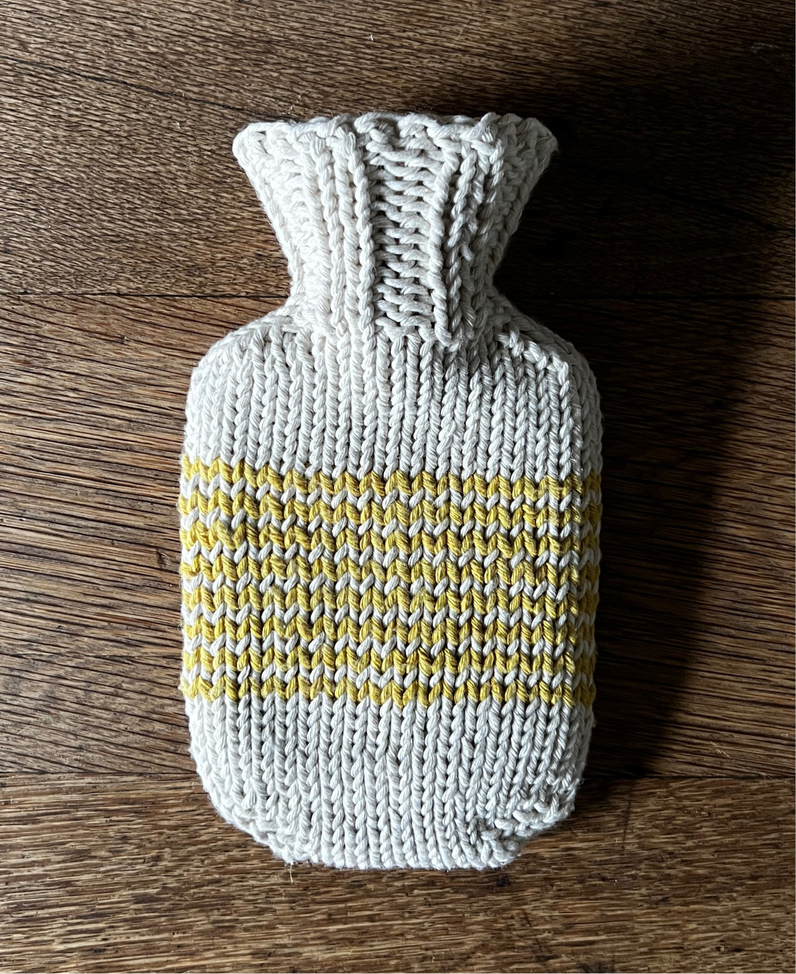 Knitted hot water bottle cover with a yellow stripe on a wooden surface