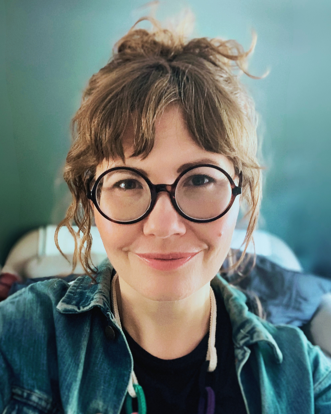 Polly Benson, founder of TTiS and Strinsel, wearing glasses and a denim jacket against a greenish blurred background