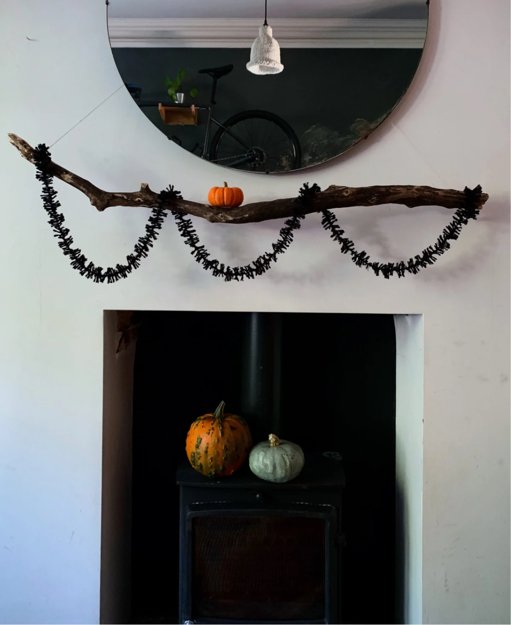 Black jute spider leg Strinsel string tinsel garland hanging from a branch suspended above a black log burner decorated with pumpkins against a white wall