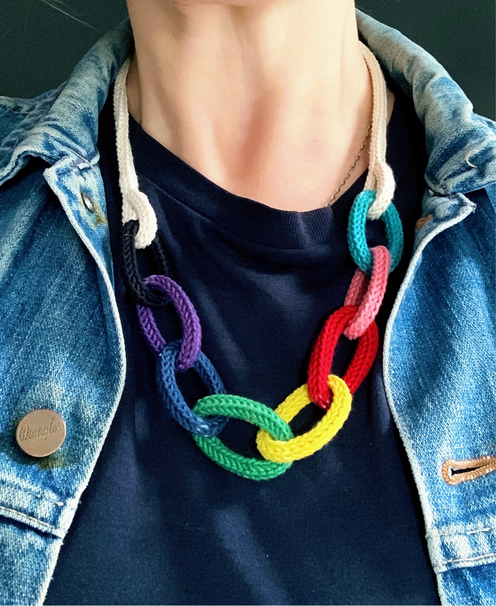 Person wearing a colorful rainbow necklace with interlocking rings, denim jacket, and dark shirt.